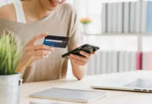Idosa olhando fatura e segurando cartões diante de mesa, com agência bancária desfocada ao fundo